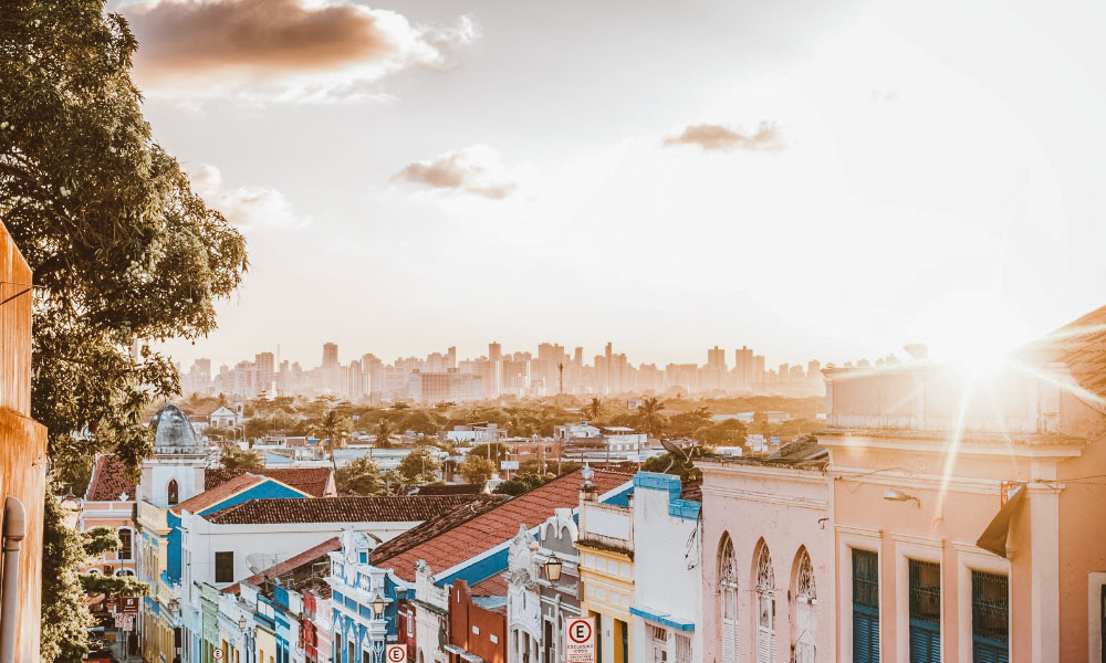 Rua com casas coloridas de Olinda e o sol se pondo sobre prédios ao fundo